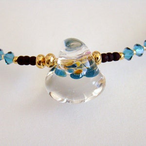 Op de afbeelding: Een ketting met een hanger van helder glas met een blauw en geel bloemenmotief. De ketting is gemaakt met kleine blauwe kristallen kralen en gouden kralen.