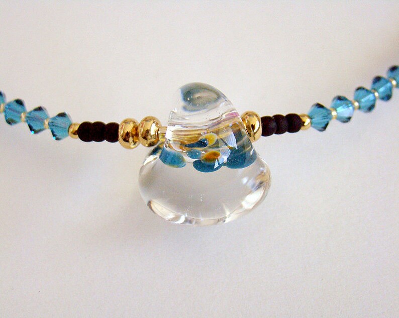 Op de afbeelding: Een ketting met een hanger van helder glas met een blauw en geel bloemenmotief. De ketting is gemaakt met kleine blauwe kristallen kralen en gouden kralen.