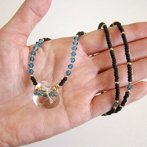 Op de afbeelding: Een ketting met een heldere glazen hanger met een libellenontwerp. De ketting is gemaakt met zwarte en gouden kralen en blauwe kristallen accenten.