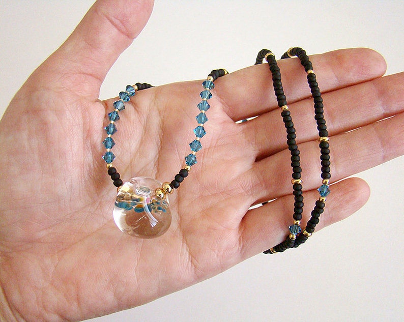 Op de afbeelding: Een ketting met een heldere glazen hanger met een libellenontwerp. De ketting is gemaakt met zwarte en gouden kralen en blauwe kristallen accenten.