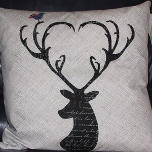 May include: A decorative throw pillow with a black deer silhouette design. The deer's antlers form a heart shape, and a small bird perches on top. The pillow has a light beige background with a textured appearance.