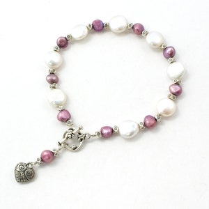 May include: A pearl bracelet featuring large, white, and smaller, purple pearls. The bracelet has a silver clasp and a heart-shaped charm. The pearls are strung together with small silver beads. The bracelet is on a white background.