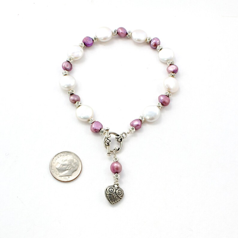 Op de afbeelding: Een parelarmband met grote, witte en kleine, paarse parels. De armband heeft een zilveren toggle sluiting en een bungelende hartbedel. De parels worden afgewisseld met kleine, heldere kralen.