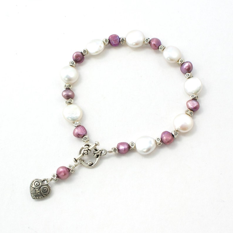 May include: A pearl bracelet featuring large, white, and smaller, purple pearls. The bracelet has a silver clasp and a heart-shaped charm. The pearls are strung together with small silver beads. The bracelet is on a white background.