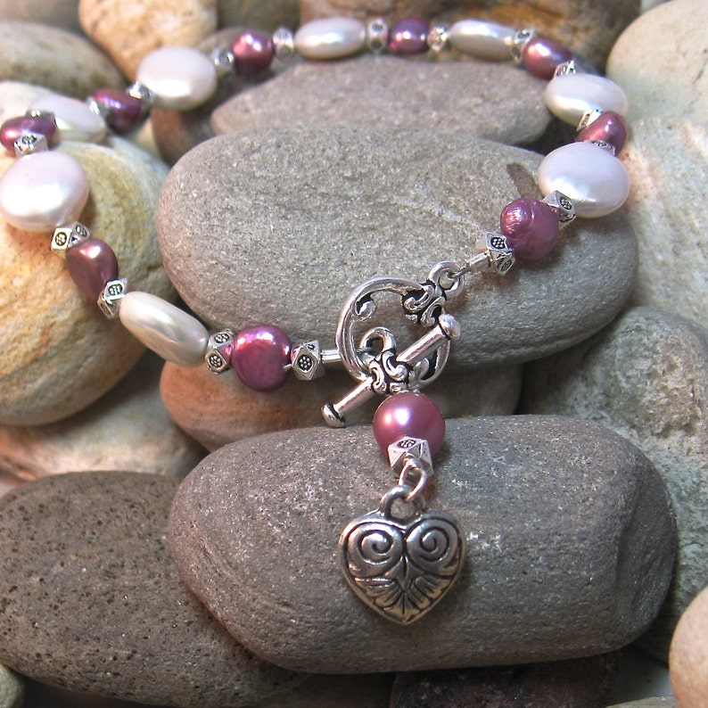 Op de afbeelding: Een ketting met een tuimelsluiting en een hartvormige hanger. De ketting is samengesteld uit witte, roze en zilveren kralen. De kralen zijn een mix van ronde en platte vormen. De ketting wordt gepresenteerd op een bed van grijze stenen.
