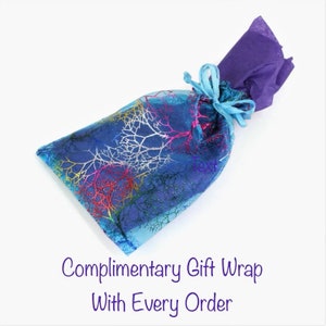 Op de afbeelding: Een blauwe cadeauzak met een boomtakontwerp in zilver, rood en geel. De zak is vastgebonden met een lichtblauw lint en bevat paars vloeipapier. De tekst "Complimentary Gift Wrap With Every Order" staat eronder.