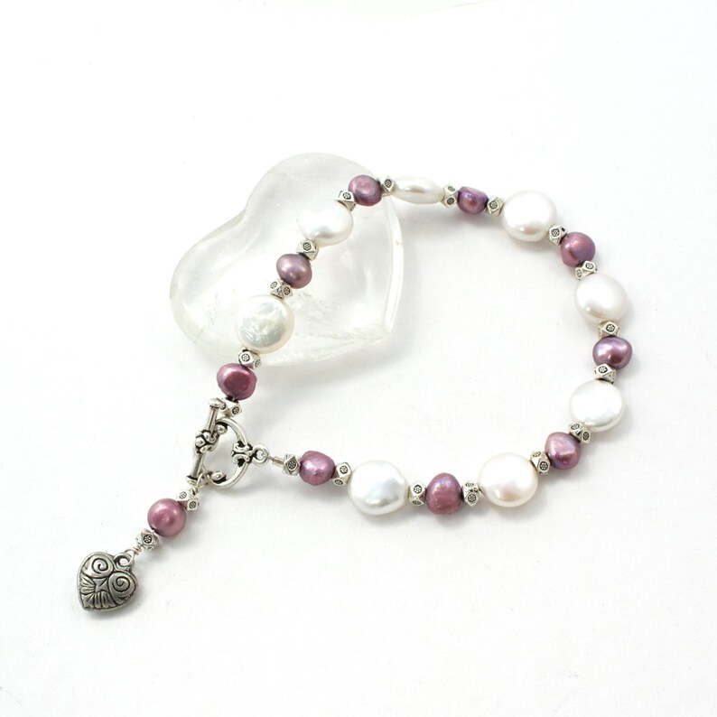 Op de afbeelding: Een parelarmband met witte en mauvekleurige parels, afgewisseld met kleinere kralen. De armband heeft een zilverkleurige toggle sluiting en een hartvormige bedel. De parels zijn van verschillende maten en vormen.