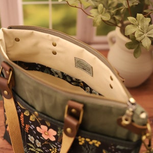 Puede incluir: Una bolsa de lona verde con un forro de estampado floral y un asa de cuero. La bolsa tiene una cremallera y un peque&ntilde;o bolsillo en la parte delantera. La bolsa est&aacute; etiquetada con "Everyday" en el interior.