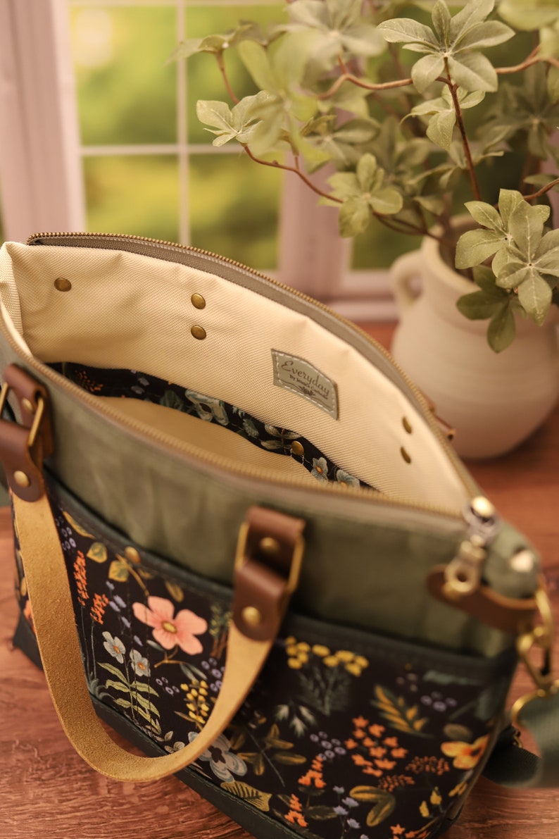 Puede incluir: Una bolsa de lona verde con un forro de estampado floral y un asa de cuero. La bolsa tiene una cremallera y un peque&ntilde;o bolsillo en la parte delantera. La bolsa est&aacute; etiquetada con "Everyday" en el interior.