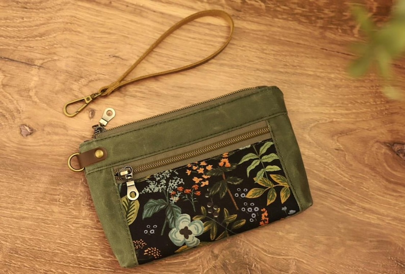 Puede incluir: Una bolsa de lona verde con cremallera y forro de estampado floral. La bolsa tiene una correa de cuero marr&oacute;n con un cierre de metal.