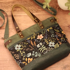 Puede incluir: Un bolso tote de lona verde con estampado floral y correas de cuero marr&oacute;n. El bolso tiene cierre de cremallera y una correa larga para llevarlo al hombro.
