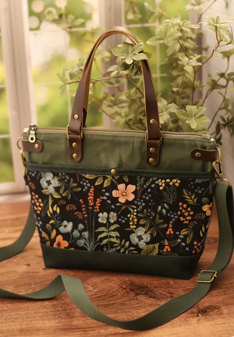 Puede incluir: Un bolso tote de lona verde con estampado floral y asas de cuero marr&oacute;n. El bolso tiene cierre de cremallera y una correa de hombro desmontable.