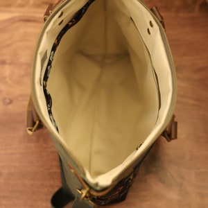 Puede incluir: Un bolso tote de lona verde con cierre de cremallera y una correa larga ajustable. El bolso tiene un forro beige y un peque&ntilde;o bolsillo interior.