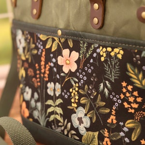 Puede incluir: Un bolso tote de lona verde con un forro de estampado floral. El bolso tiene asas de cuero marr&oacute;n y cierre de cremallera. El bolso tambi&eacute;n tiene una correa larga verde.