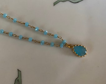 Könnte beinhalten: Eine zarte goldene Kettenkette mit hellblauen Glasperlen und einem Anhänger mit einem hellblauen Edelstein, der in einen goldenen Rahmen gefasst ist.