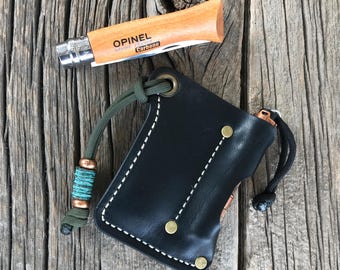May include: A black leather knife sheath with a white stitch detail and a green cord with a turquoise bead. The sheath has a brass snap closure and is designed to hold a small pocket knife. The knife is not included.