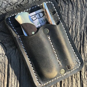 May include: A black leather wallet with white stitching, holding a pen, a small knife, and a US $50 bill. The wallet has a snap closure and the word "USA" stamped on the bottom right corner. The wallet is on a weathered wooden surface.