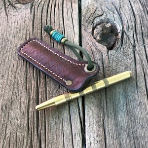 May include: A brown leather sheath with a white stitch detail and a green cord with a turquoise bead. A gold metal tool with a textured surface is lying on top of the sheath.