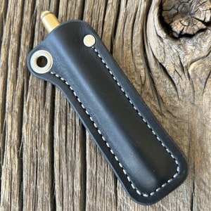 May include: A black leather pen case with white stitching and a brass top. The case has a metal loop on the side.