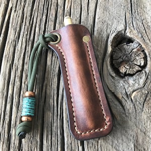 May include: A brown leather sheath with a green paracord lanyard and a turquoise bead. The sheath has a brass snap closure and is designed to hold a small tool or pen.