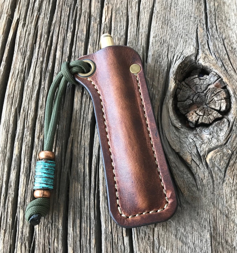 May include: A brown leather sheath with a green paracord lanyard and a turquoise bead. The sheath has a brass snap closure and is designed to hold a small tool or pen.