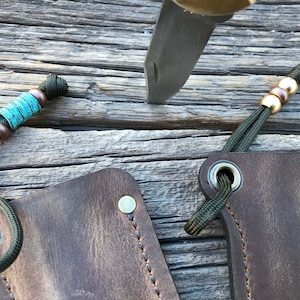 May include: Two brown leather knife sheaths with green paracord lanyards and metal accents. One sheath has a turquoise and copper bead on the lanyard, the other has two copper beads.