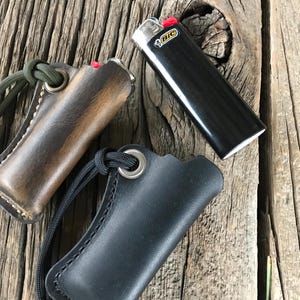 May include: Two leather cases for lighters, one brown and one black, with a black lighter with the word "BIC" on it. The cases have a loop for attaching to a keychain.