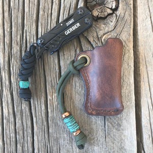 May include: A brown leather multi-tool sheath with a green paracord lanyard and a turquoise bead. The sheath has a metal ring at the top. A black Gerber Dime multi-tool is attached to the lanyard.