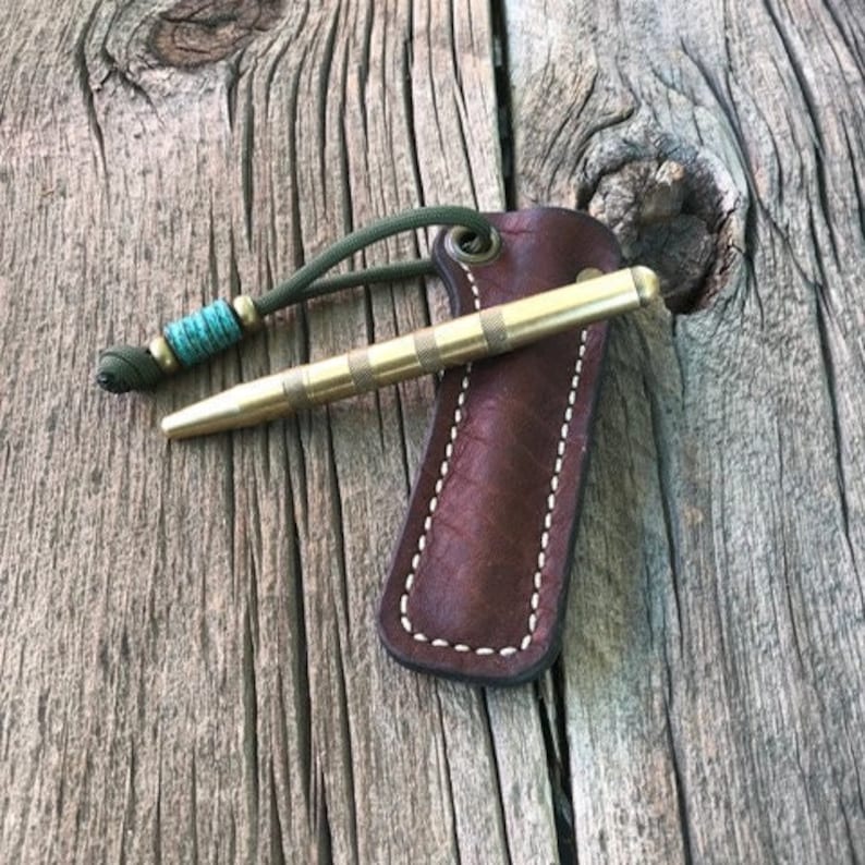 May include: A brass pen with a brown leather sheath and a green and blue paracord lanyard. The pen is lying on a wooden surface.