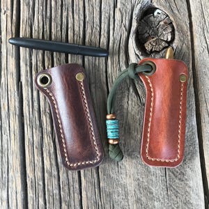 May include: Two leather sheaths for a pen or small knife. The sheaths are brown and have a stitched edge. One sheath has a green cord with a turquoise bead attached.