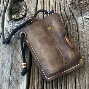 May include: A brown leather pouch with a drawstring closure and a stitched design. The pouch has two pockets and is designed to hold small items like a knife or a flashlight.