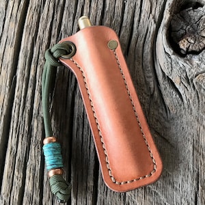 May include: A brown leather sheath with a stitched edge and a green cord with a turquoise bead. The sheath is designed to hold a small, cylindrical object.