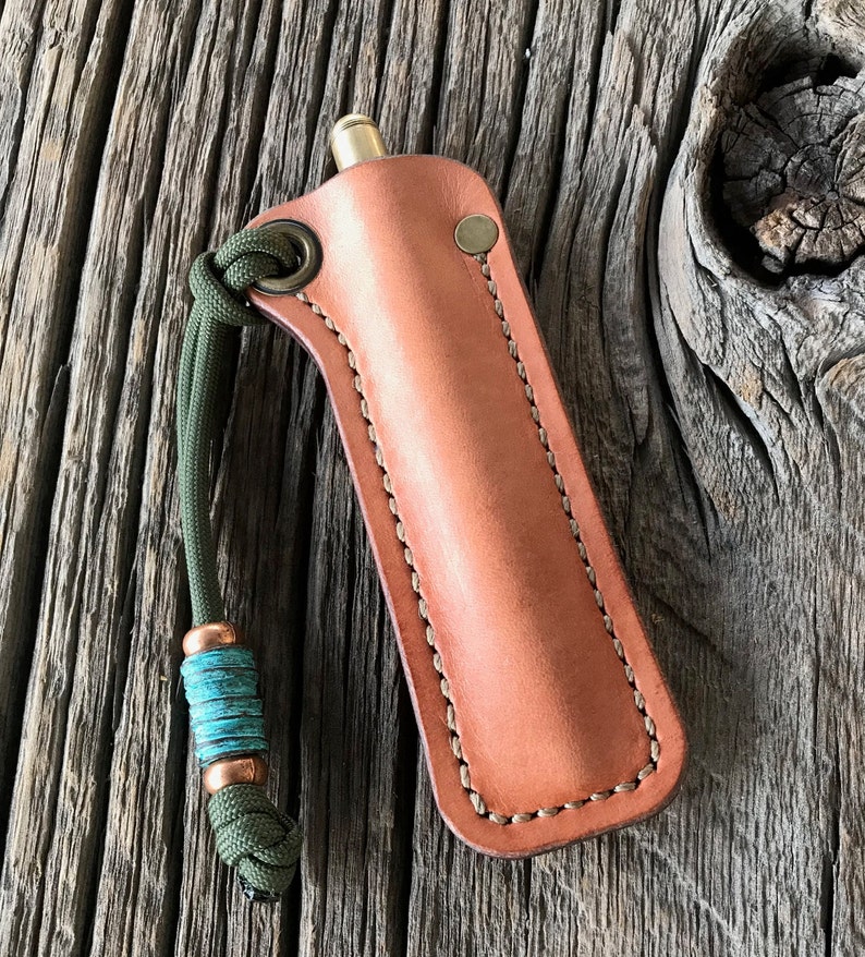 May include: A brown leather sheath with a stitched edge and a green cord with a turquoise bead. The sheath is designed to hold a small, cylindrical object.
