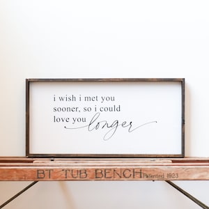 May include: A framed sign with a white background and black text that reads "i wish i met you sooner, so i could love you longer". The sign is sitting on a wooden bench.
