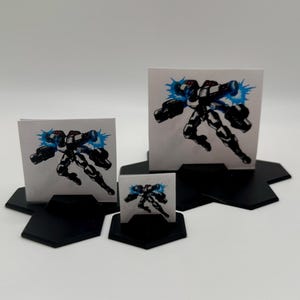 Puede incluir: Tres piezas de juego de papel impresas de diferentes tamaños que representan un robot blanco y negro con explosiones de energía azules. Las piezas están sobre bases hexagonales negras.