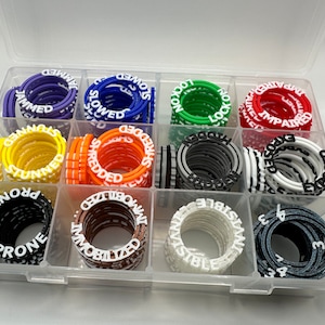 Puede incluir: Una caja de almacenamiento de plástico transparente con 12 compartimentos que contienen pilas de anillos de plástico de colores. Cada anillo tiene una palabra diferente impresa en él, como "atascado", "ralentizado", "desgarrado", "deteriorado", "expuesto" y "propensos".