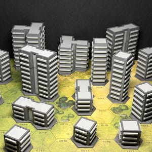 May include: A collection of miniature grey and white plastic building models arranged on a hexagonal grid game board. The structures feature varied heights and rectangular designs with horizontal window detailing, representing a city landscape.