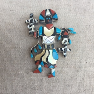 May include: A colorful, hand-crafted, Native American-style pin depicting a stylized figure with turquoise, coral, and abalone shell accents.