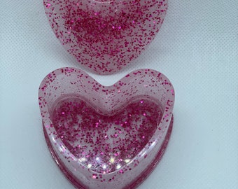May include: Two heart-shaped trinket boxes. The boxes are clear with pink glitter embedded throughout. The top box is a lid, and the bottom box is a container. The boxes are on a white background.