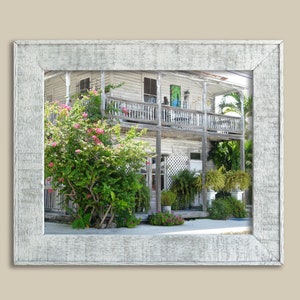 Puede incluir: Un edificio de madera blanca y desgastado con un balcón y flores rosas en frente. El edificio tiene una valla blanca de madera y un césped verde.