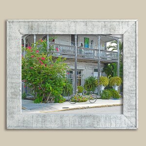 Puede incluir: Un edificio de madera blanca y desgastado con un porche y una bicicleta estacionada en frente. El edificio tiene un mural colorido en el lateral y exuberantes plantas verdes que crecen a su alrededor.