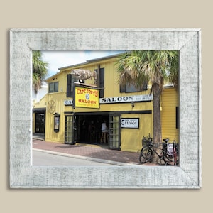 Könnte beinhalten: Ein gelbes Gebäude mit einem Schild, auf dem "Capt. Tony's Saloon" steht, und einer großen Fischskulptur, die über dem Eingang hängt. Vor dem Gebäude steht ein Palmenbaum und zwei Fahrräder sind davor geparkt.