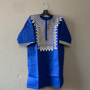 May include: A blue tunic with white embroidered details. The tunic has short sleeves and a decorative pattern around the neckline and down the front. The garment is hanging on a black hanger against a neutral wall.