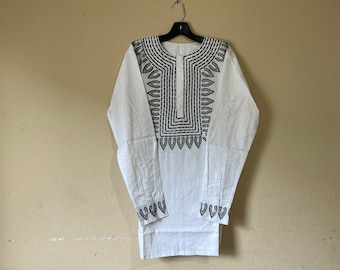 May include: White long-sleeve shirt with intricate black embroidery around the neckline, down the front, and on the cuffs. The shirt is hanging on a black hanger against a neutral wall. The embroidery features geometric and floral patterns.