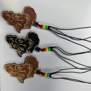 May include: Three wooden pendants in the shape of Africa, each with a different finish: light brown, dark brown, and black. Each pendant has a black cord with red, yellow, and green beads. The pendants are displayed on a white surface.