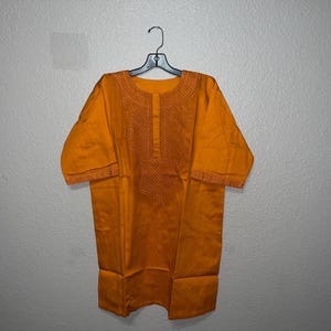 May include: Mustard yellow tunic with short sleeves, hanging on a black hanger. The tunic features embroidered detailing around the neckline and sleeves. The fabric appears to be a lightweight material.