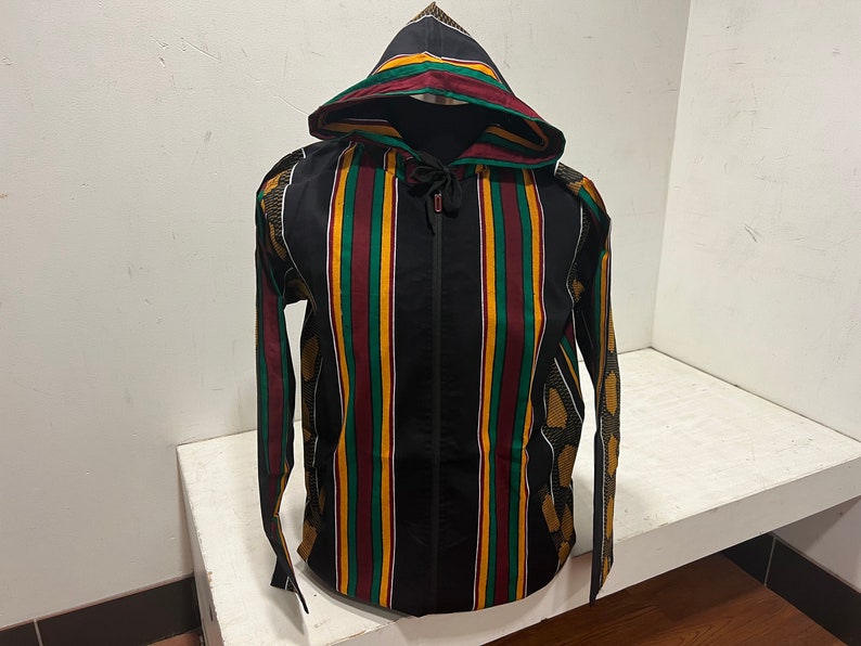 Pu&ograve; includere: Una giacca nera con cappuccio e chiusura a zip. La giacca ha strisce verticali in rosso, verde e giallo.