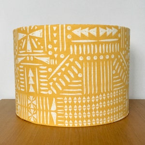 May include: A cylindrical drum lampshade featuring a mustard yellow background with a repeating white geometric pattern consisting of triangles, vertical lines, dots, and dashes arranged in a structured, block-like composition across the surface.