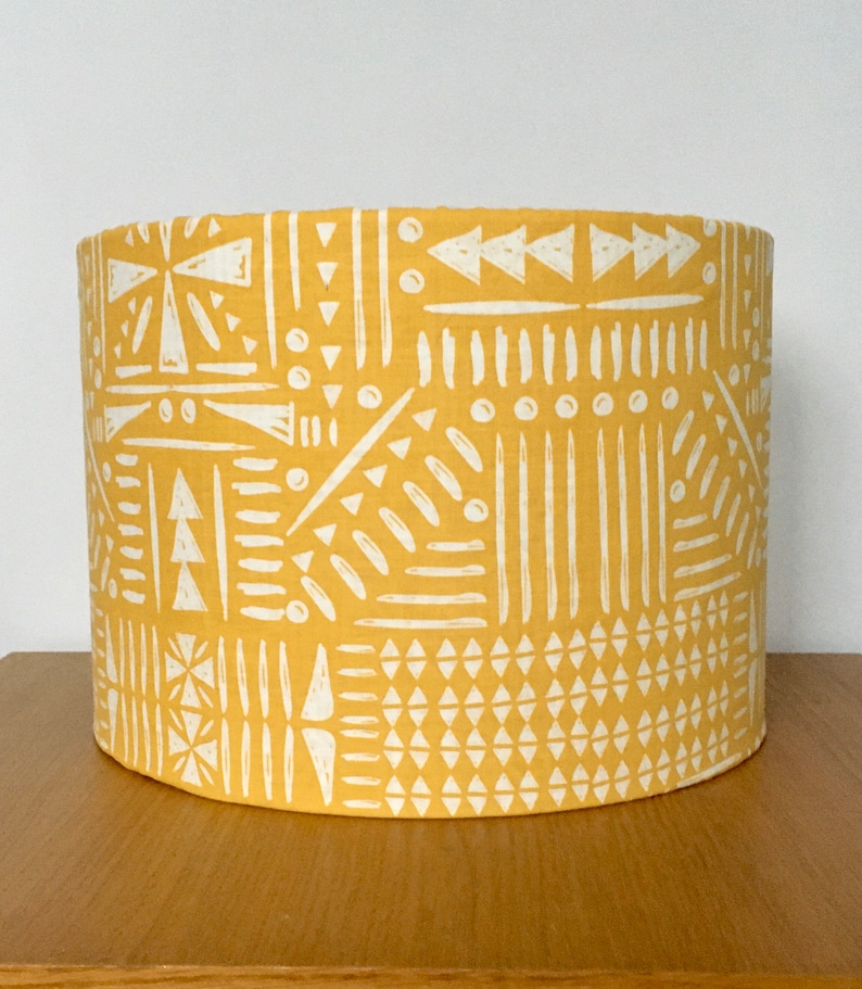 May include: A cylindrical drum lampshade featuring a mustard yellow background with a repeating white geometric pattern consisting of triangles, vertical lines, dots, and dashes arranged in a structured, block-like composition across the surface.