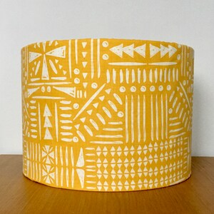 May include: A cylindrical lampshade featuring a vibrant mustard yellow background with a repeating white geometric pattern. The design consists of various hand-drawn style shapes including triangles, vertical lines, dots, and dashes arranged in panels.